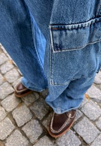 Salopette in denim blu chiaro con gambe larghe e tasca frontale, abbinata a scarpe in pelle marrone scuro con cuciture a contrasto.