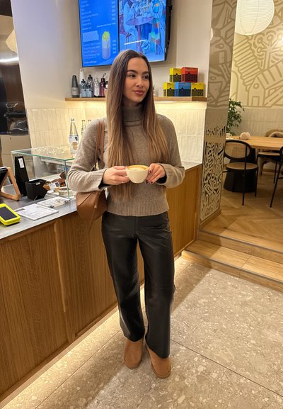 Joven mujer con cabello largo sostiene una taza de café dentro de un café, de pie cerca del mostrador con pantallas de menú y asientos en el fondo.
