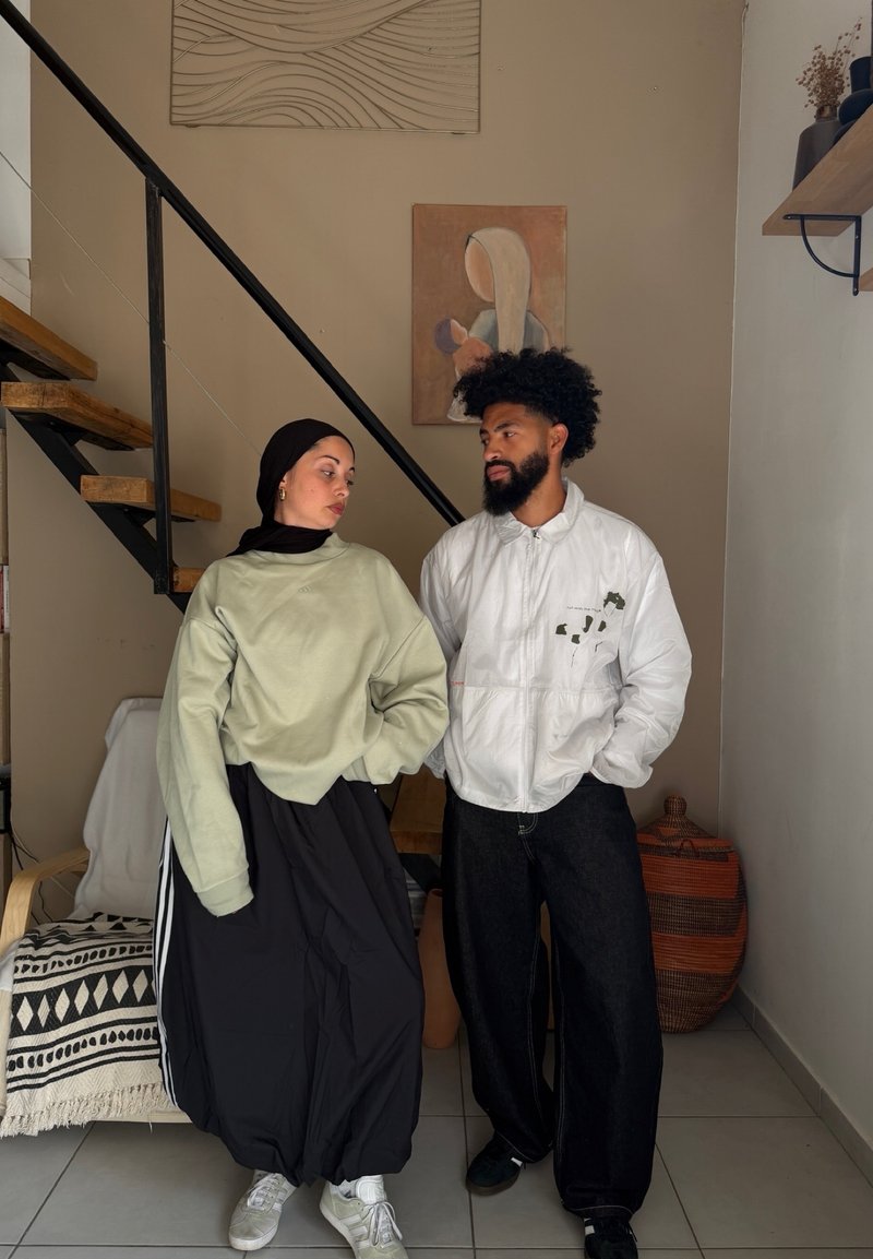 Un homme et une femme se tiennent à l'intérieur près des escaliers, face à face ; la femme porte un hijab et des vêtements amples, l'homme porte une veste blanche et un pantalon noir.