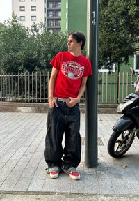 Piros póló, amelyen a "Bajnokok" felirat látható, laza sötét farmernadrággal, fehér övvel és piros sportcipővel, zöldellő városi háttér előtt.