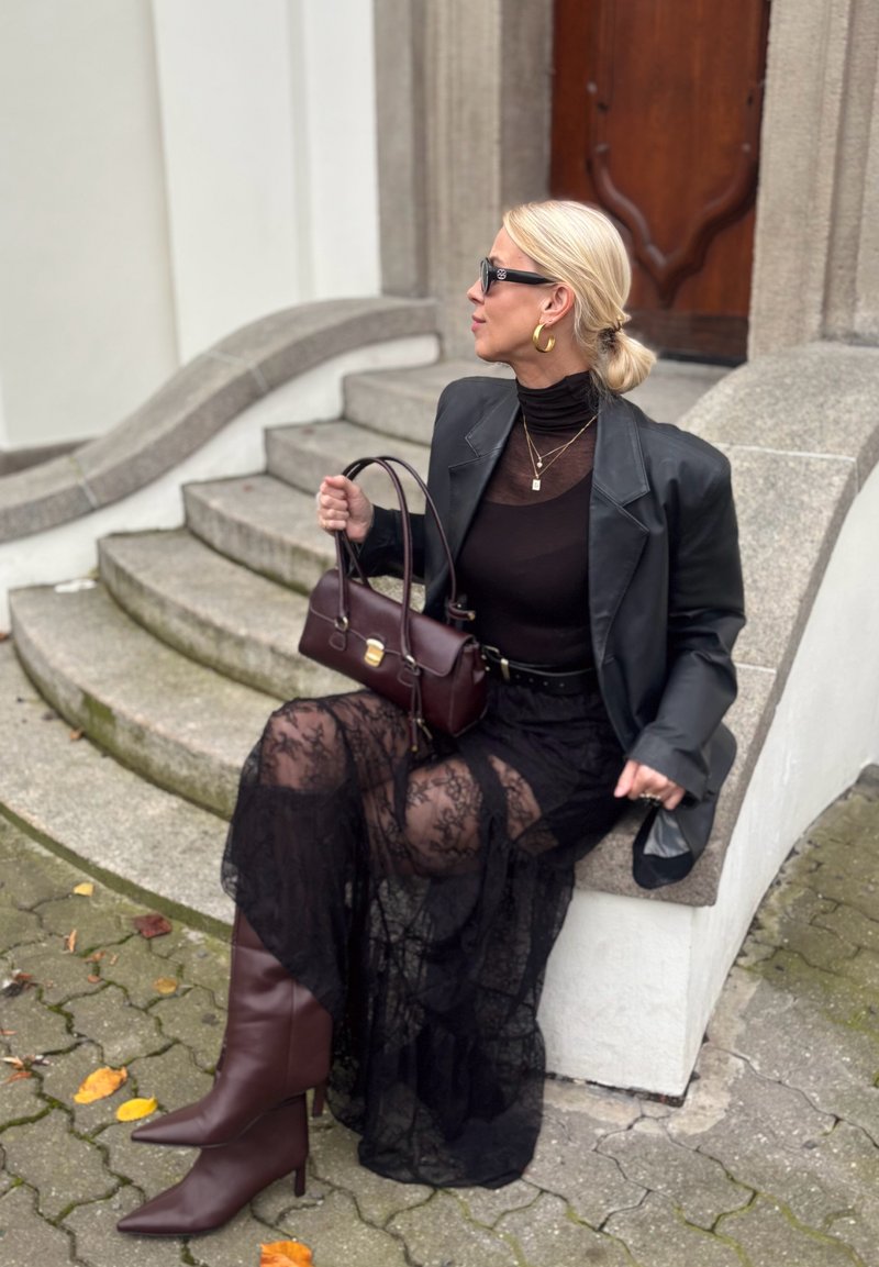Black lace dress layered over a sheer top, paired with a structured black blazer. Brown knee-high boots and a burgundy handbag in hand.