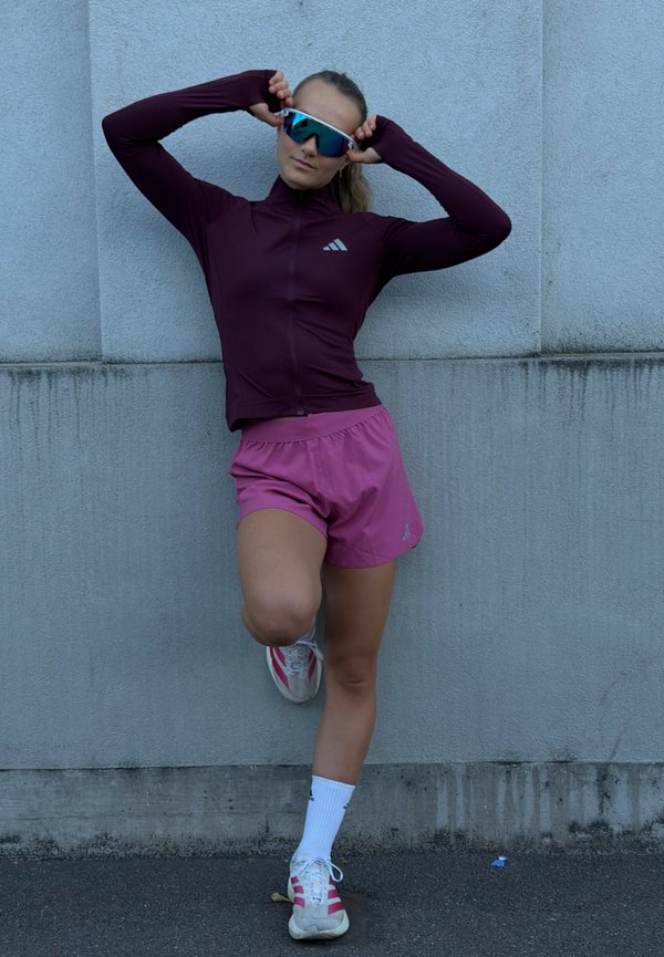 Une femme en tenue de course ajuste ses lunettes de soleil à verres réfléchissants tout en étirant une jambe contre un mur en béton.