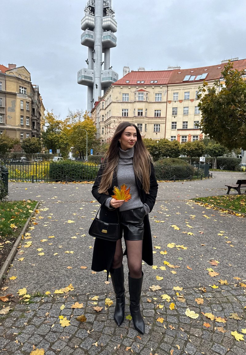 Mladá žena v čiernom kabáte, šedom svetri, kožených šortkách a čižmách drží oranžové jesenné listy v mestskom parku s vysokou vežou a budovami v pozadí.