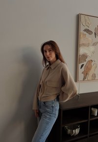 Pikkade pruunide juustega naine, kes kannab beežikat jopet ja siniseid teksaseid, seisab tumeda puidust kapikese ja abstraktse seinakunsti lähedal neutraalses ruumis.