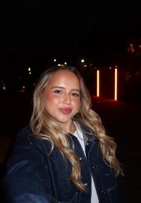 Giacca di jeans di colore blu scuro con bottoni argentati, indossata sopra una maglietta bianca. Sullo sfondo, illuminazione soffusa e accenti al neon.