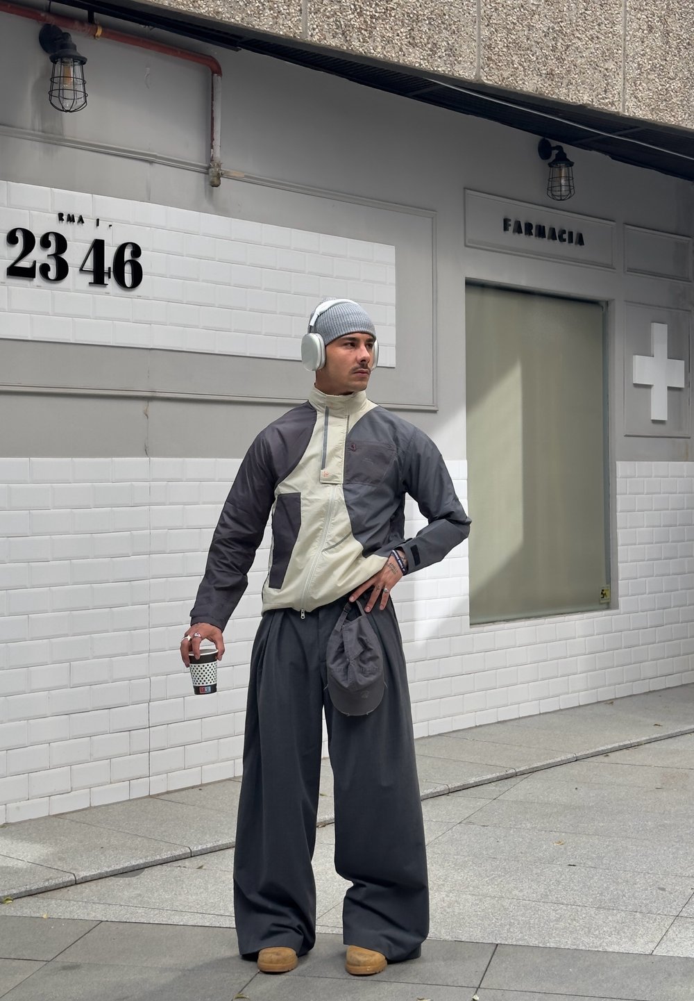Leichte Jacke in Creme und Dunkelgrau, lockere dunkelgraue Hose, graue Mütze, Kopfhörer, hält einen Kaffeebecher, mit einer lässigen Tasche.