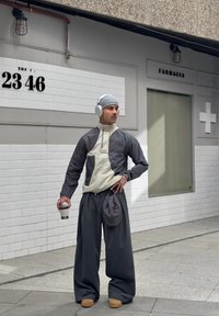 Leichte Jacke in Creme und Dunkelgrau, lockere dunkelgraue Hose, graue Mütze, Kopfhörer, hält einen Kaffeebecher, mit einer lässigen Tasche.