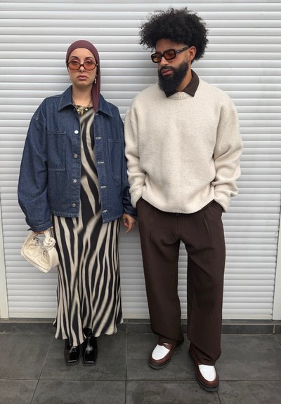Dos personas están de pie frente a una persiana blanca; una lleva un vestido con rayas de cebra, chaqueta vaquera, pañuelo en la cabeza, gafas de sol y sostiene un bolso; la otra lleva un suéter crema, pantalones marrones y zapatos bicolor.