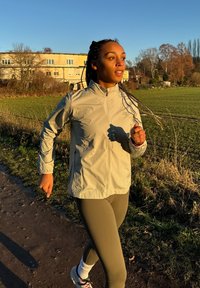 Veste de course beige clair à col montant, fermeture éclair et logo discret, associée à des leggings vert olive et des baskets blanches, présentée en plein air.