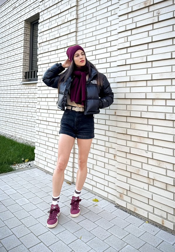 Schwarze Pufferjacke, beiges Oberteil, dunkle Jeans Shorts; bordeauxroter Beanie und Schal; weiße Sneaker mit bordeauxroten Akzenten; grauer Pflasterboden, Backsteinwand im Hintergrund.