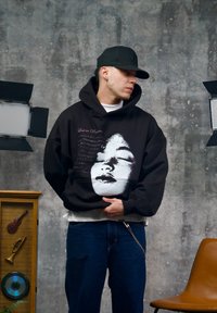 Sweat à capuche noir avec un graphisme représentant un visage et du texte en blanc et violet. Le modèle porte un jean foncé et un sous-pull blanc.