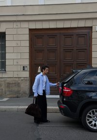 Muž s veľkou hnedou taškou otvára kufor čierneho SUV zaparkovaného pri obrubníku pred budovou s veľkými drevenými dverami.