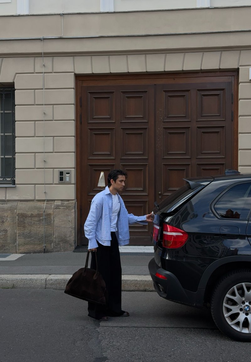 Homem com uma grande bolsa castanha a abrir a mala de um SUV preto estacionado junto ao passeio em frente a um edifício com grandes portas de madeira.