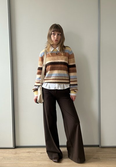 Joven mujer de pie en el interior contra una pared lisa, vistiendo un suéter de rayas, camisa blanca con cuello, pantalones anchos de rayas finas y un bolso de hombro.