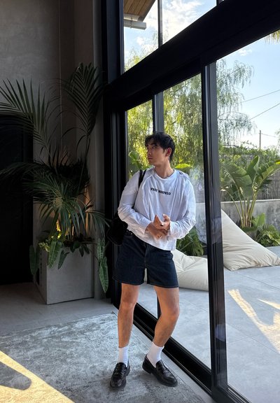 Camisa blanca de manga larga con texto negro, pantalones cortos de mezclilla oscura, zapatos negros y calcetines blancos. Ambiente interior con grandes ventanas y plantas.
