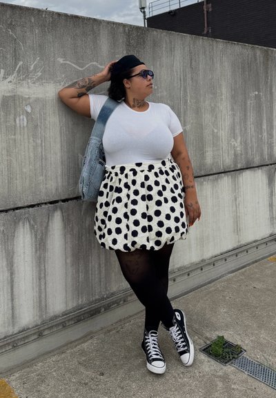 Camiseta blanca, falda de lunares en blanco y negro, medias negras y zapatillas Converse negras. Lleva un bolso de mezclilla y gafas de sol.