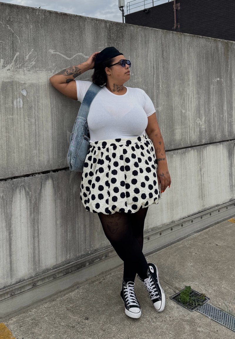 Camiseta branca, saia de bolinhas brancas e pretas, collants pretos e ténis Converse pretos. Usa uma mala de ganga e óculos de sol.