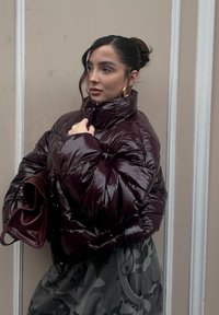Burgundy padded jacket with a glossy, quilted texture, high collar, and zip, paired with camo-patterned trousers and a burgundy handbag.
