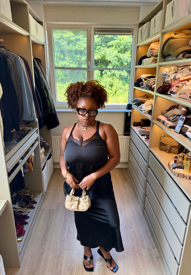 Femme aux cheveux bouclés et portant des lunettes, vêtue d'une robe noire tenant un petit sac beige, debout dans un dressing avec des vêtements et des chaussures.