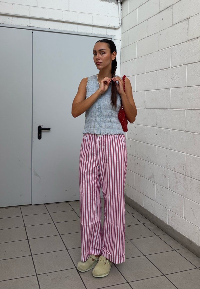 Jeune femme tressant ses cheveux portant un haut gris sans manches, un pantalon rayé rouge et blanc, des chaussures beige et tenant un sac rouge.
