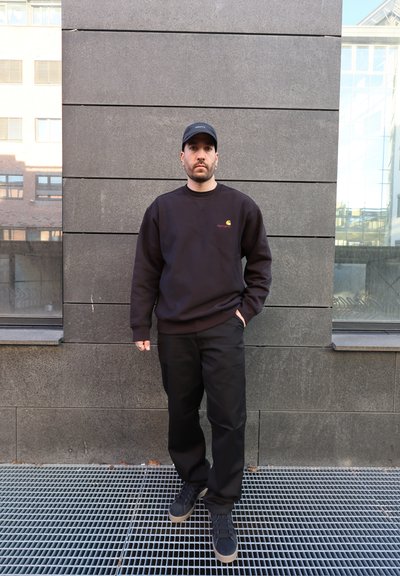 Hombre con sudadera negra Carhartt, pantalones negros y gorra negra, de pie contra una pared de azulejos grises con ventanas a ambos lados.
