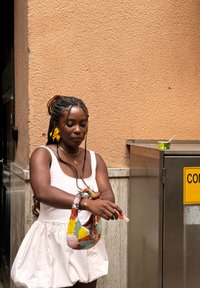 Donna in abito bianco con capelli intrecciati e fiore giallo applica gel igienizzante per le mani vicino a un contenitore di metallo con una tazza sopra.