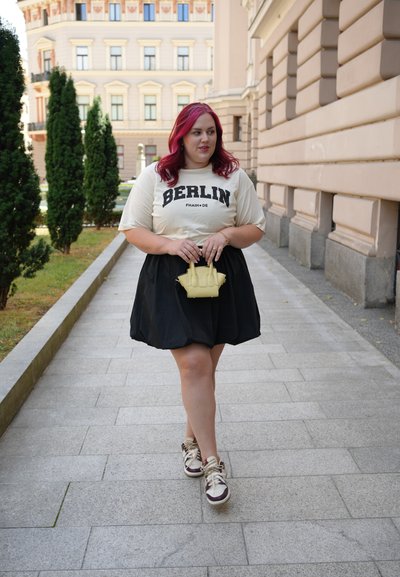 Falda negra, camiseta beige con la impresión "BERLÍN", bolso amarillo y zapatillas en tonos neutros. El cabello es de longitud hasta los hombros y de color rojizo-pinken.