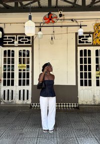 Top senza spalline in denim, pantaloni bianchi in lino e una borsa nera. Lo sfondo presenta porte di legno, luci appese e un ventilatore da soffitto rosso.