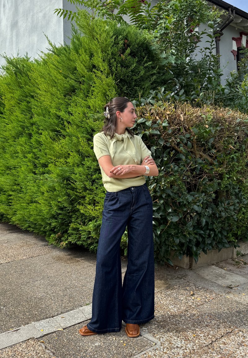 Blusa verde de manga curta com detalhe de nó, combinada com jeans de perna larga em denim escuro e sandálias castanhas. O fundo apresenta vegetação e pavement.