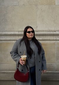 Casaco cinza de botão duplo, suéter cinza, jeans azuis, segurando uma bebida gelada em um copo transparente e uma bolsa bordeaux. Óculos de sol retangulares usados.