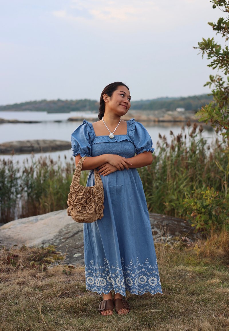 Woman in a blue dress with puffed sleeves holding a woven bag, standing on grass near water and rocks with trees in the background.