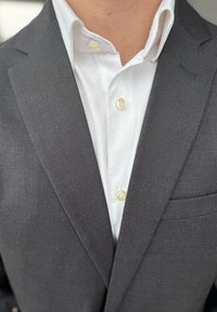 Grå kostymjacka med notch-lapel, en vit skjorta med krage under och ljusa knappar. Inkluderar en bröstfickdetalj.