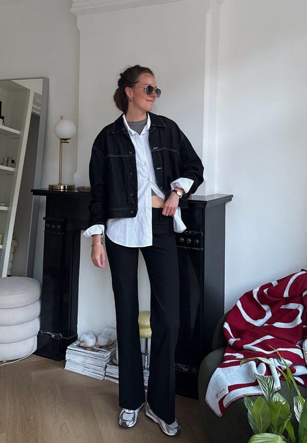 Ženska z očali za sonce, v črni denim jakni čez belo majico in sivo zgornjo majico, črnih hlačah in supergah, stoji v sodobni sobi z odprtim kaminom.