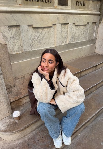 Joven mujer con chaqueta de forro polar blanca y vaqueros azules sentada en unos escalones de piedra junto a una taza de café, mirando pensativa frente a una pared de mármol.