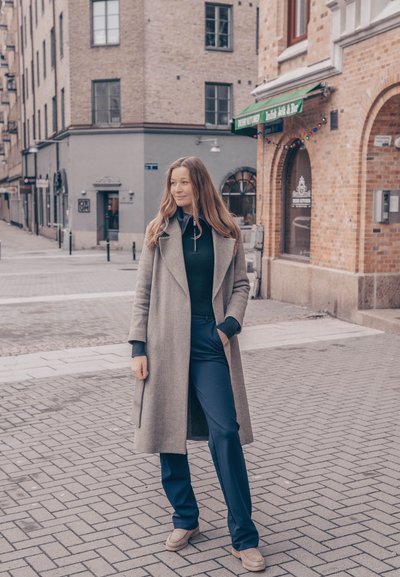 Abrigo largo gris sobre un suéter negro de cuello alto, combinado con pantalones azul marino y zapatos beige; edificios de ladrillo texturizado de fondo con pavimento limpio.