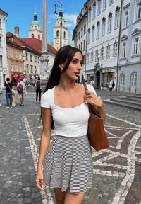 Λευκό ριγέ τοπ, ριγέ φούστα, καφέ τσάντα; λιθόστρωτος δρόμος, ιστορικά κτίρια και μια πλατεία στο φόντο κάτω από έναν γαλάζιο ουρανό.