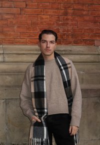 Pull beige en tricot avec un ourlet côtelé, associé à une écharpe à carreaux noire et grise ornée de franges, porté sur un pantalon foncé.