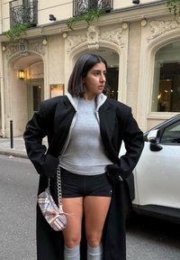 Kvinne med svart bob-frisyre iført en oversized svart kåpe, grå genser, svarte shorts, grå leggvarmere og holder en rutete veske på byens gate.