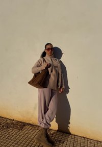 Suéter bege claro sobre uma blusa de canelado, calças rosa suave, mala de camurça castanha com elementos metálicos, e botas de amarrar bege. Sombra projetada na parede.