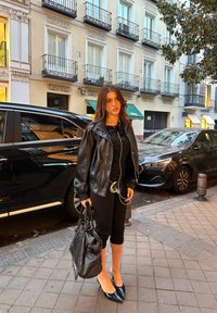 Čierna kožená bunda s lesklou textúrou, čierne tričko, čierne skrátené nohavice, špicaté čierne podpätky; nesie čiernu tašku a slúchadlá. Mestské prostredie.