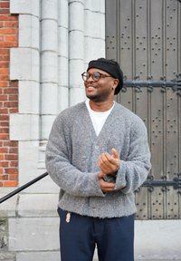 Cardigan gris et duveteux avec des boutons sur le devant, porté par-dessus une chemise blanche, associé à un pantalon bleu marine. Accessoires comprenant un béret noir et des lunettes rondes.