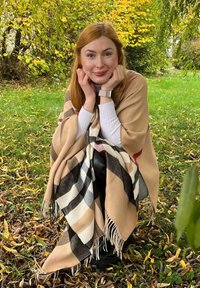 Châle à motif écossais beige et noir avec des franges, drapé sur les épaules, porté sur un haut à manches longues blanc et un pantalon noir.