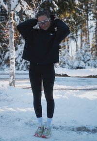 Black cropped jacket, black leggings, white socks, and multicoloured running shoes with yellow and pink accents; snowy background and trees.