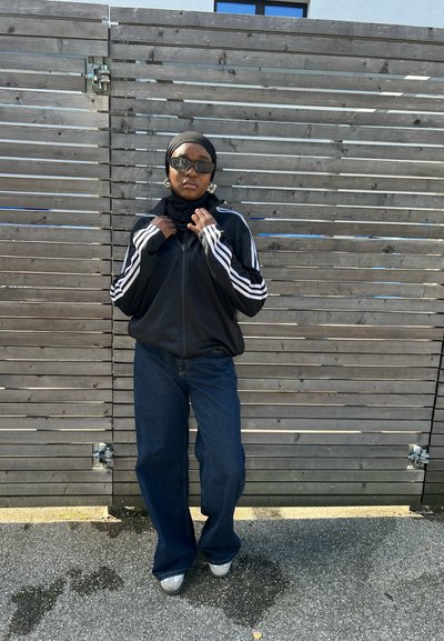 Chaqueta negra con cremallera y rayas blancas, pantalones vaqueros anchos de mezclilla oscura y gafas de sol, combinados con un hijab negro frente a un fondo de madera.