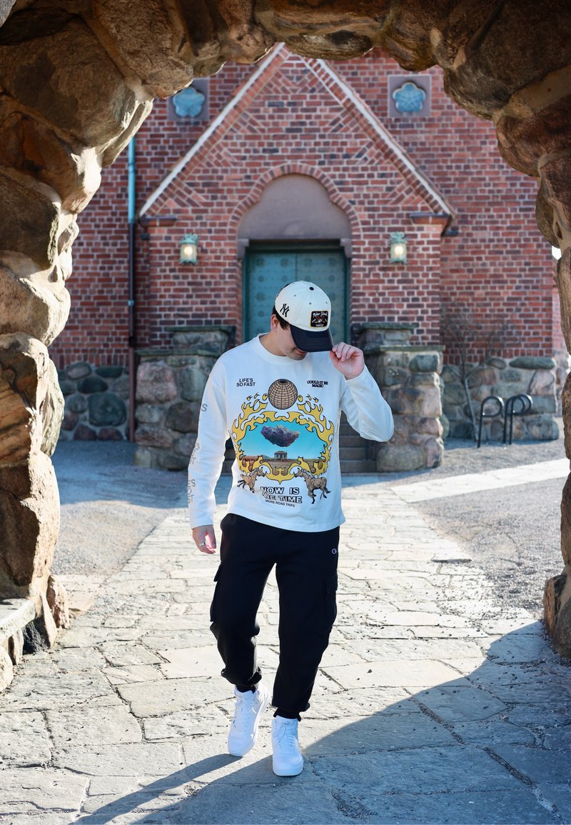 Junger Mann, der seine Mütze richtet, trägt ein Sweatshirt mit Grafikdruck und Sneakers, geht auf einem Steinweg unter einem felsigen Torbogen in der Nähe eines Backsteingebäudes.