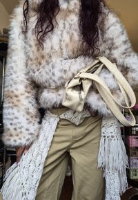 Personne portant une veste en fausse fourrure à taches, un pantalon beige, tenant un sac à main crème avec une longue bandoulière et une écharpe blanche à franges crochetées.