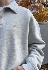 Tecido de pulôver cinza claro com acabamento texturizado, apresentando uma gola, dois botões e um pequeno logótipo bordado a ouro no peito.