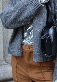 Sivá pletená cardigan s rebrovanými manžetami, strieborný top s flitrami a hnedé zamšové nohavice s dekoračným šitím a čierna kožená crossbody taška.