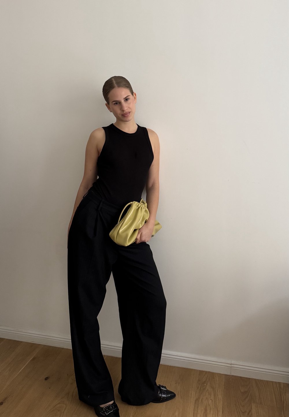 Czarna bezrękawnikowa bluzka, czarne spodnie o wysokim stanie, trzymająca miękką, żółtą skórzaną torebkę. Gładkie materiały i luźny, szeroki krój.