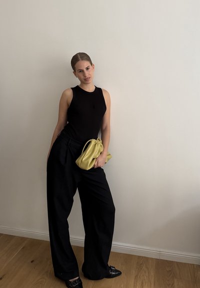 Top negro sin mangas, pantalones negros de talle alto, sosteniendo un suave clutch de cuero amarillo. Texturas suaves y ajuste holgado y relajado.